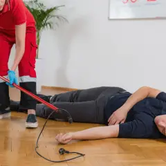 Ratownik medyczny w czerwonym stroju zabezpiecza miejsce wypadku, gdzie leży mężczyzna po porażeniu prądem. W tle tablica z napisem "CPR".