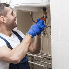 Instalator w niebieskich rękawiczkach pracuje przy bojlerze, przygotowując się do tego, jak spuścić wodę z bojlera.