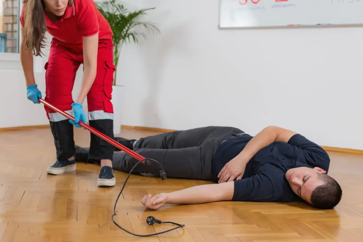 Ratownik medyczny w czerwonym stroju zabezpiecza miejsce wypadku, gdzie leży mężczyzna po porażeniu prądem. W tle tablica z napisem "CPR".