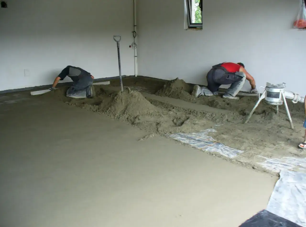 Dwóch robotników układa półsuchy beton na wylewkę, wyrównując go łopatami i łatami.