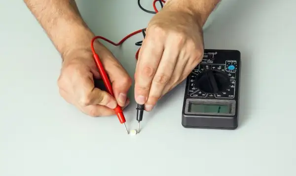 Ręce trzymające sondy multimetru dotykające małego białego elementu elektronicznego.